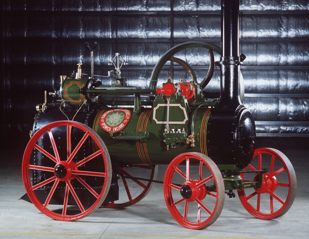 A horizontal riveted boiler with a firebox at one end mounted on four red-pained iron wheels. A flywheel is mounted on one side, a chimney at the front takes smoke away, and a cylinder, connecting rods, valves and governor are mounted on top of the boiler. The engine is finished in olive green with fine lining and a company transfer is on the side.